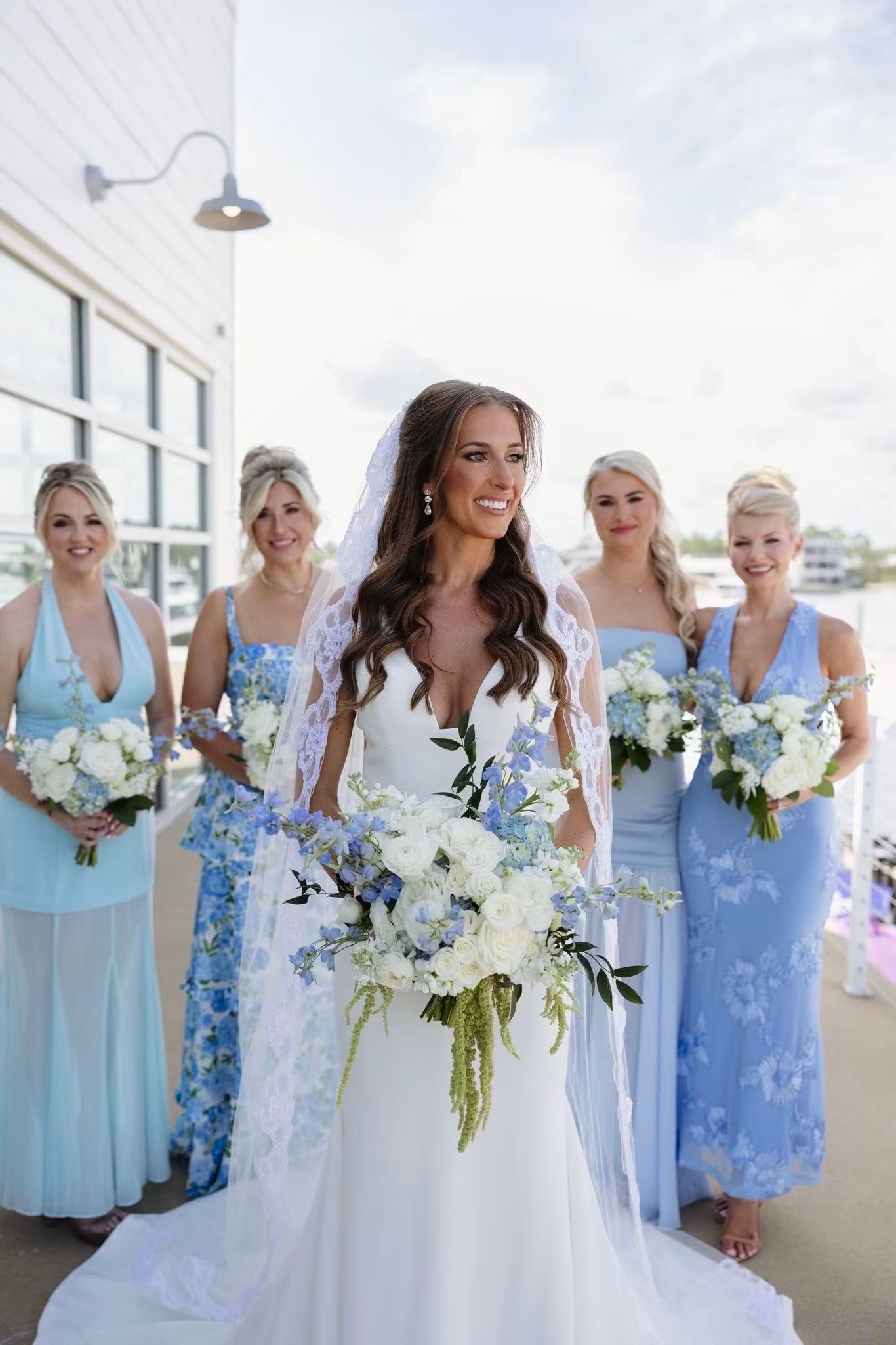 Bride in white gown holds blue and white bouquet with four bridesmaids in light blue dresses at waterfront wedding.