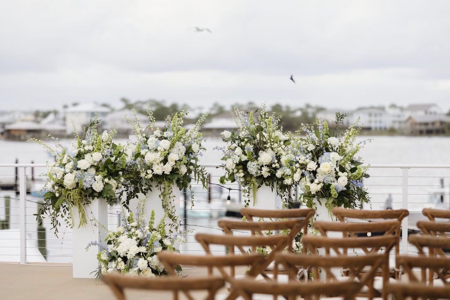 Outdoor waterfront wedding ceremony setup with white floral arrangements on tall pedestals and wooden chairs overlooking a marina.
