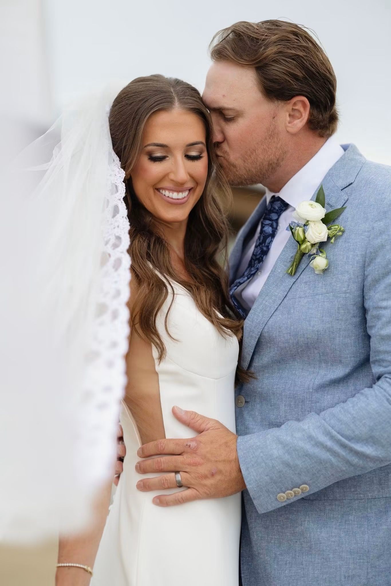 Bride in white gown and veil smiles as groom in light blue suit kisses her cheek during wedding portrait.