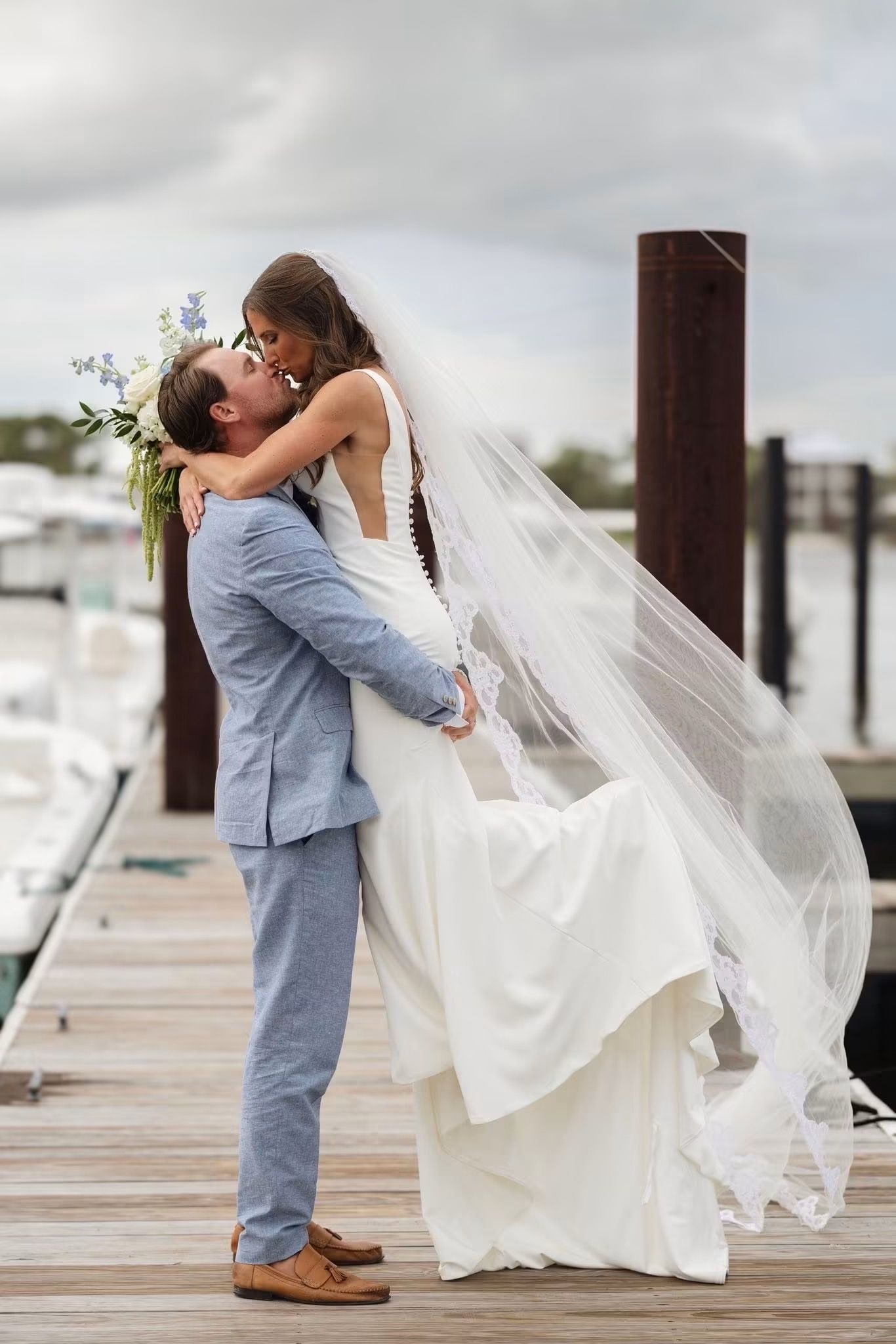Groom lifts bride in white gown and veil on a marina dock, sharing a romantic kiss on their wedding day.