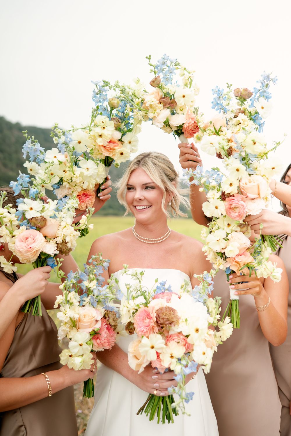 Bridesmaids in brown dresses hold colorful bouquets with white and coral flowers against a sunny outdoor backdrop.