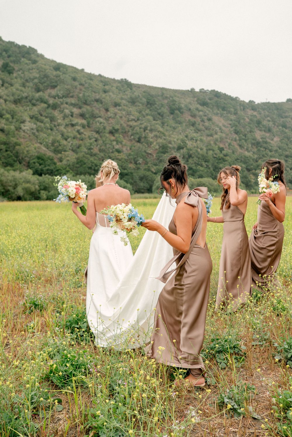 Wedding party in neutral dresses walks through a wildflower meadow with green mountains in the background.