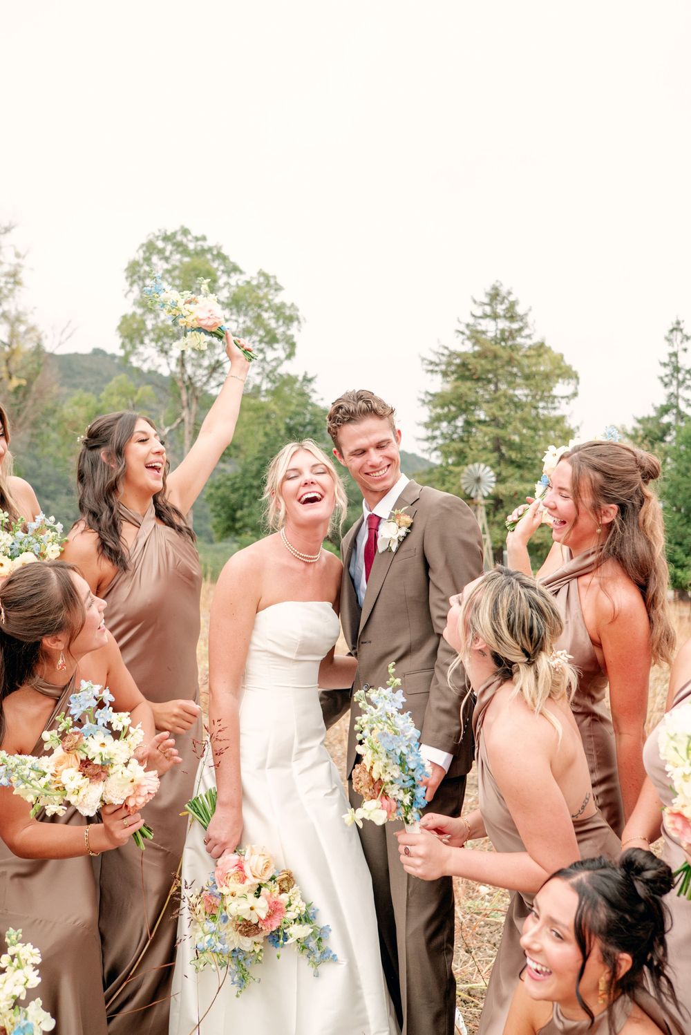 A wedding party in brown suits and pink dresses laughs joyfully together during an outdoor photo session.