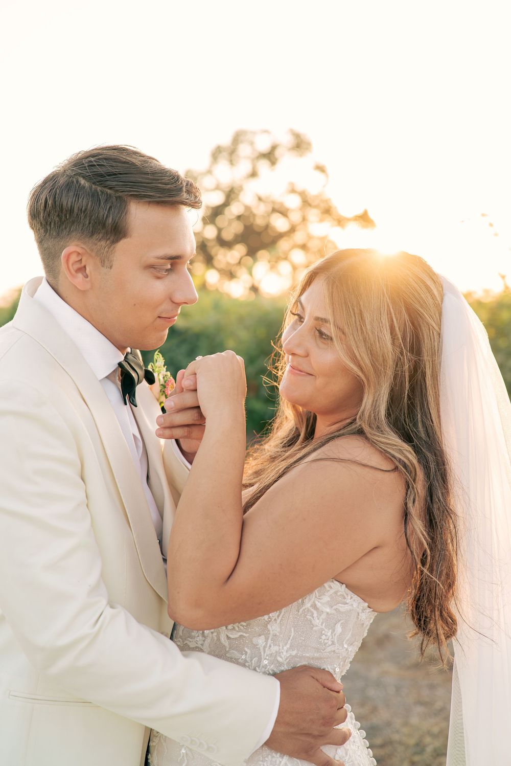 A romantic wedding couple embraces during golden hour sunset in a field wearing white attire and a veil.