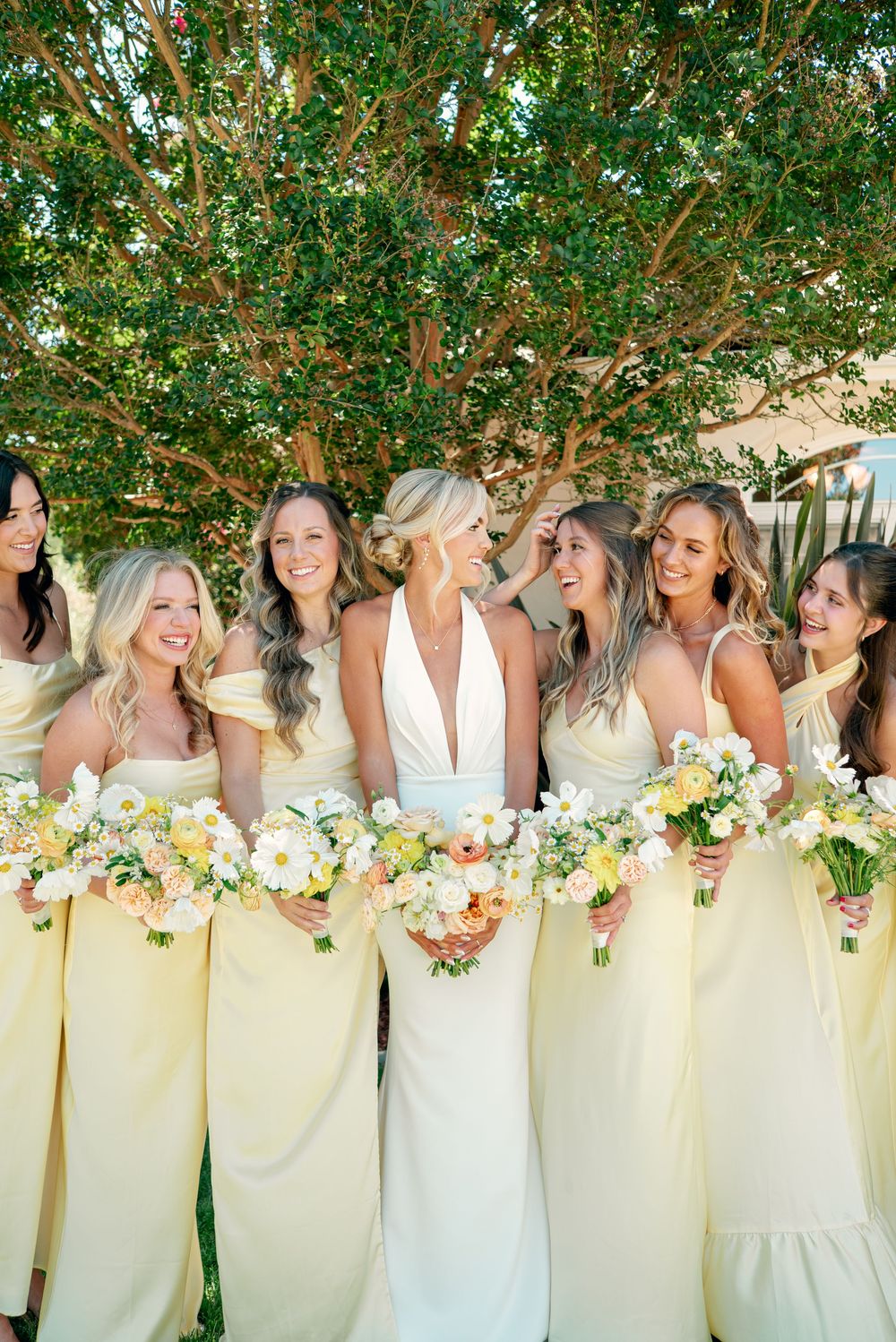 Bridesmaids in pale yellow dresses stand in a row holding white and yellow floral bouquets under green trees.