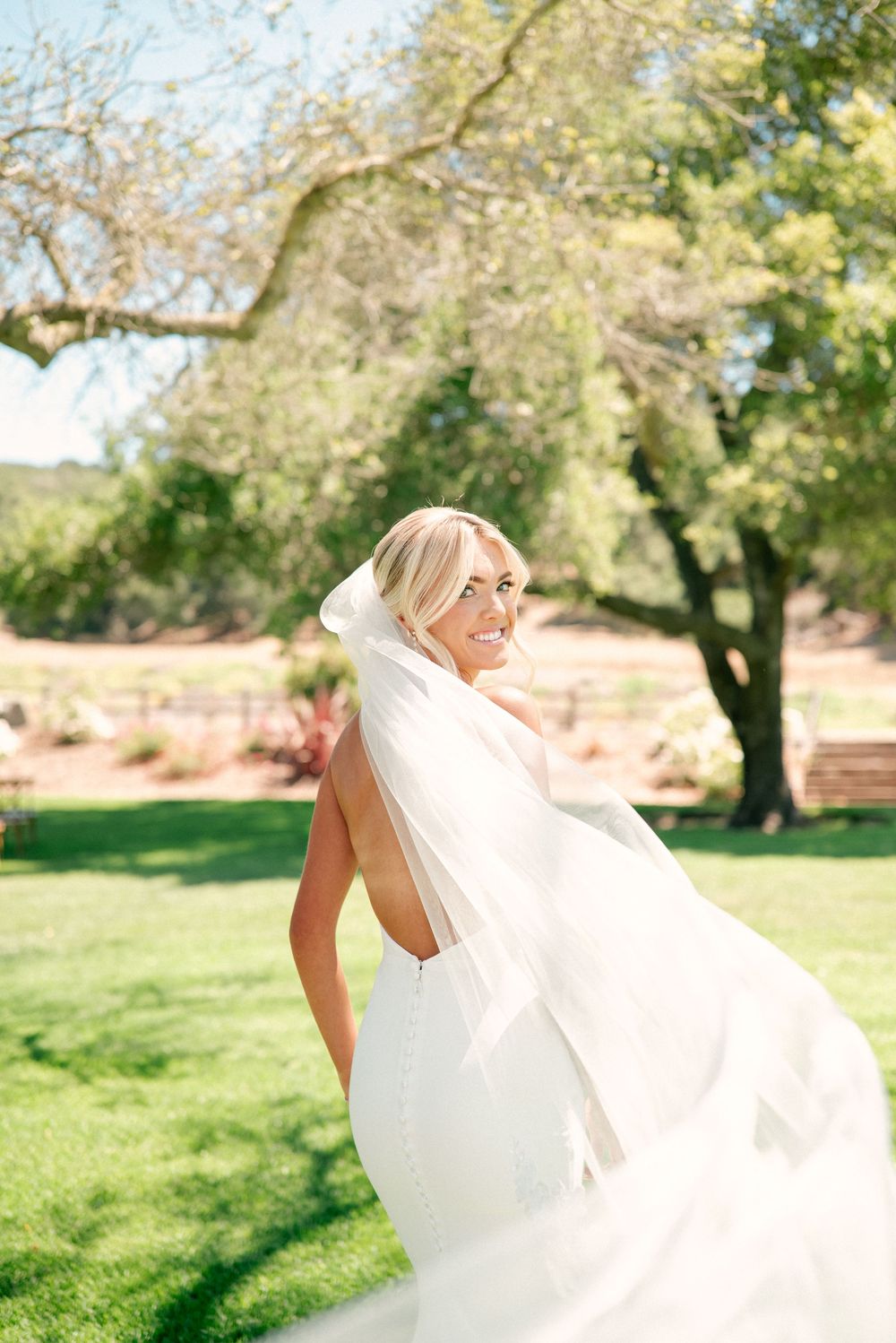 A bride in a flowing white wedding dress twirls joyfully in a sunny outdoor garden setting.