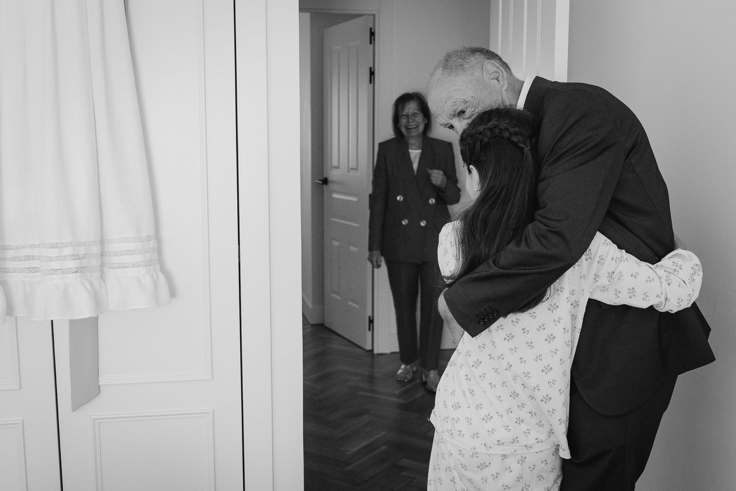 A black and white wedding photo showing an emotional embrace between people in a doorway while someone watches from behind.
