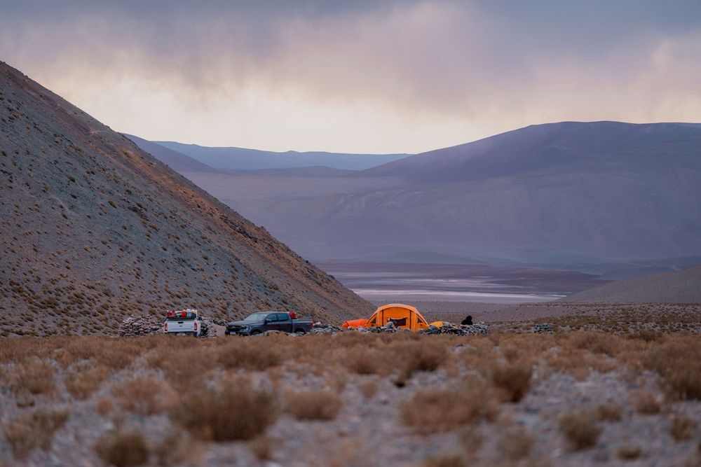 Orange tent glows at dusk among parked vehicles on a mountainside overlooking a desert valley.