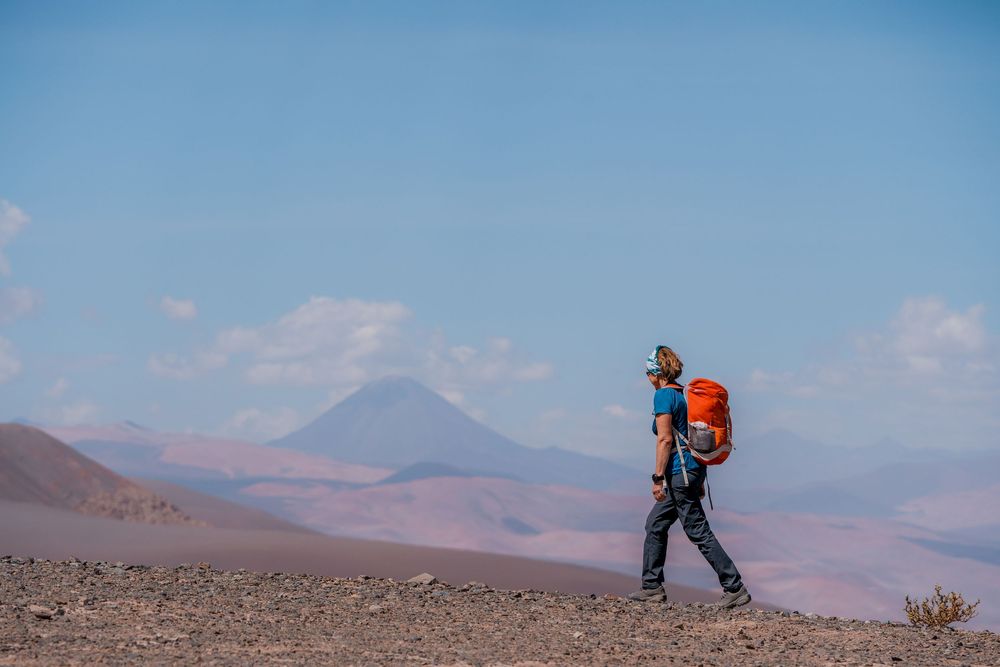 Hikers with backpacks traverse a rugged mountain trail against a backdrop of distant peaks and blue sky.