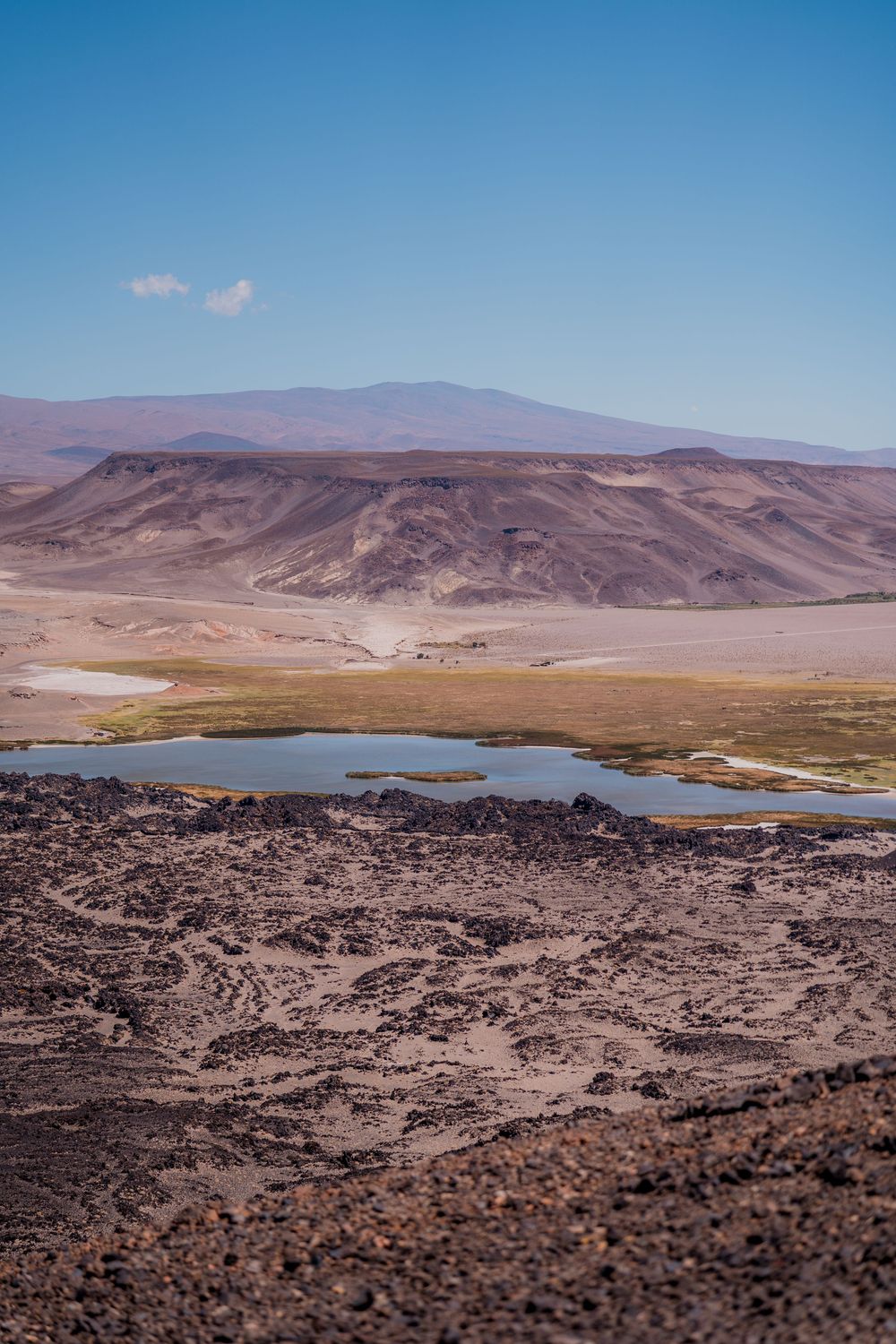 Desert mountain landscape with small lake and rocky terrain under clear blue sky in arid region.