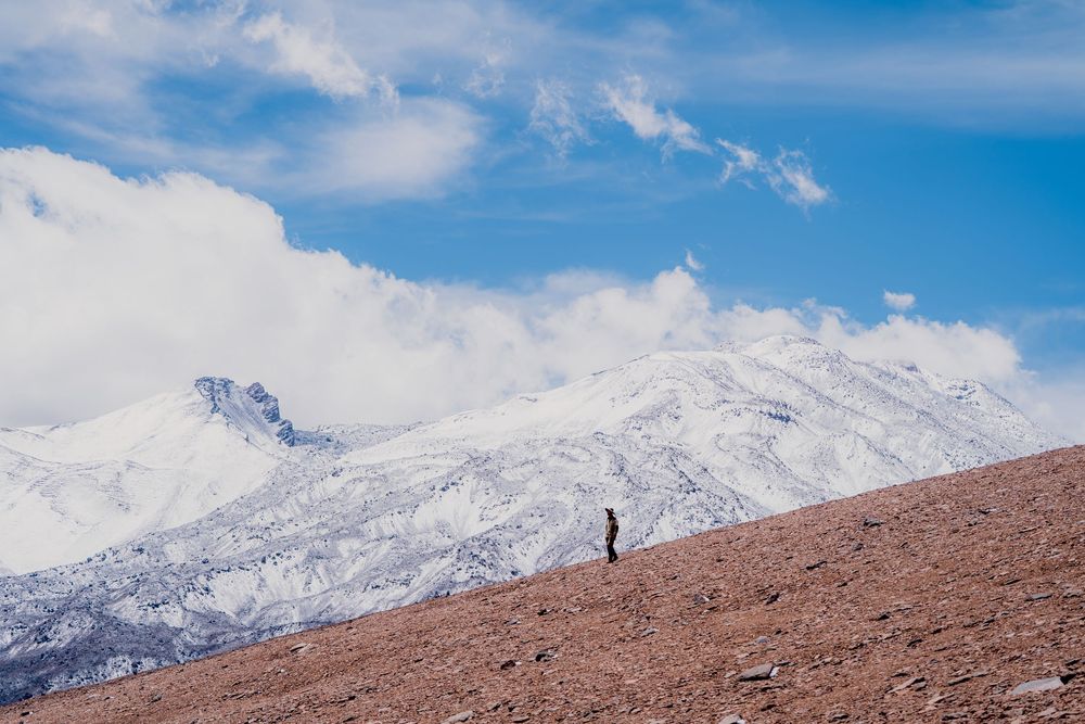 A hiker traverses a rugged mountain ridge against a backdrop of snow-capped peaks and blue skies.