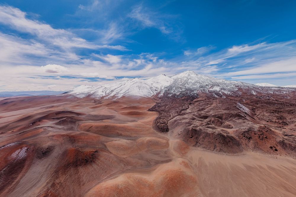 Aerial panorama of snow-capped mountains and desert landscape under bright blue sky with wispy clouds.