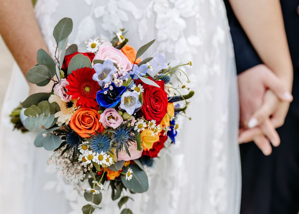 Vibrant wedding bouquet with red, blue, and orange flowers mixed with eucalyptus leaves against white dress fabric.