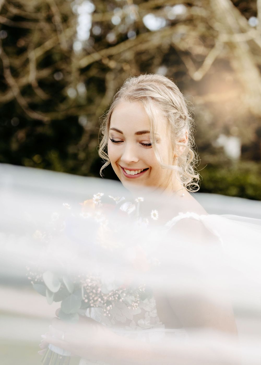 A bride in a white dress smiles joyfully through soft dreamy veil fabric on her wedding day.