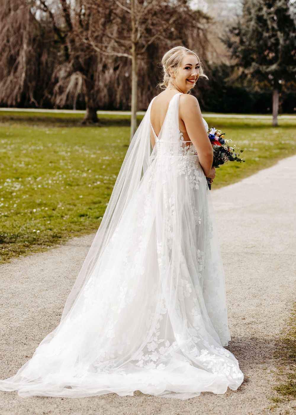 A bride in a flowing white wedding gown with a long veil walks along a garden path on a sunny day.