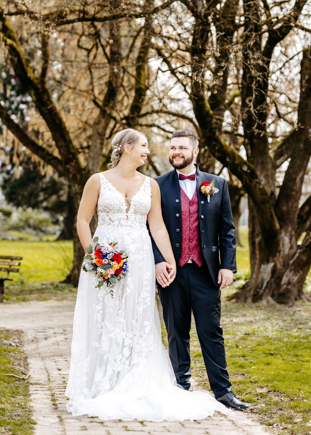 A couple in wedding attire walks along a dirt path surrounded by bare trees in a natural outdoor setting.