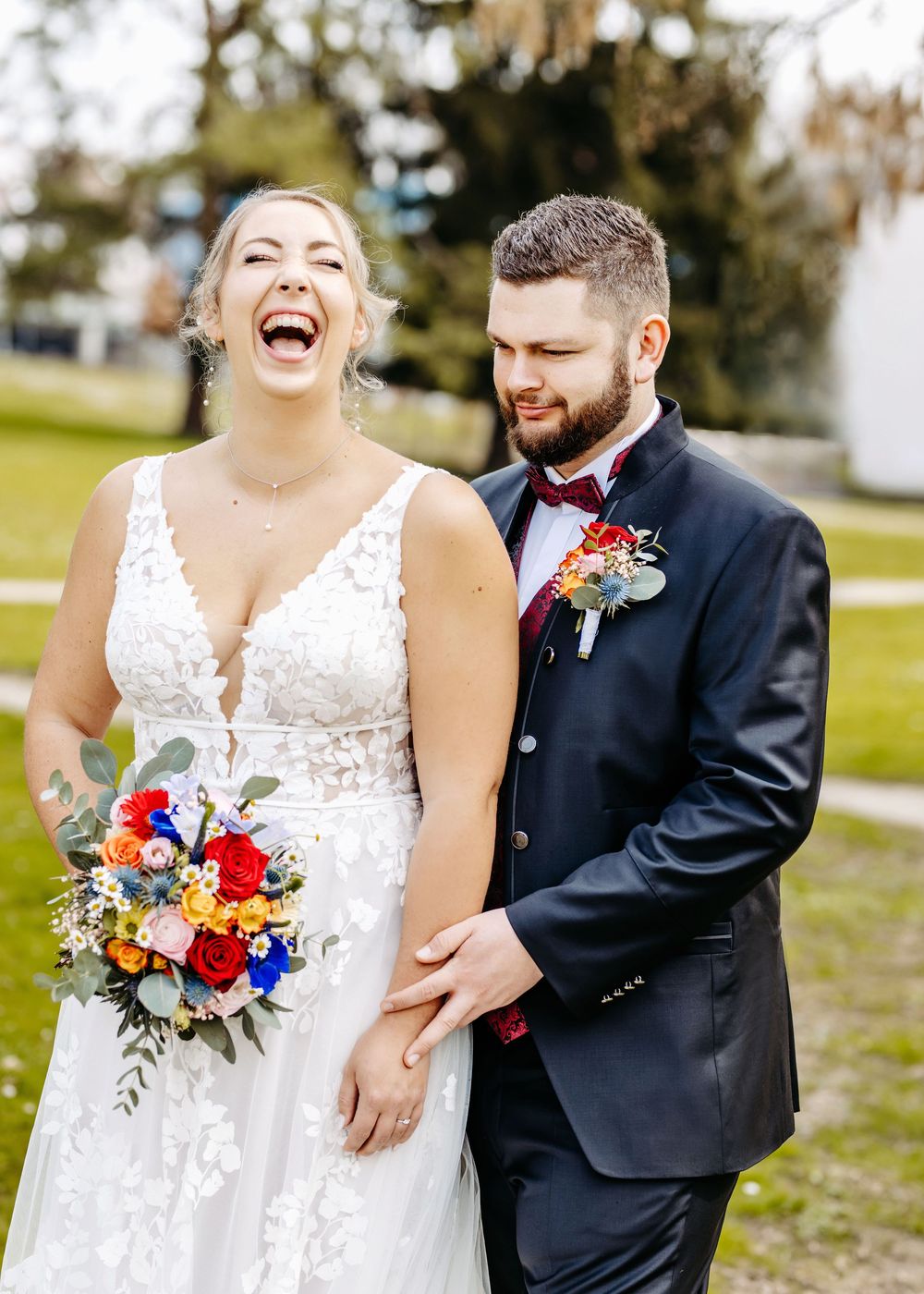 A joyful wedding couple shares a candid moment outdoors with colorful red and blue floral bouquet details.