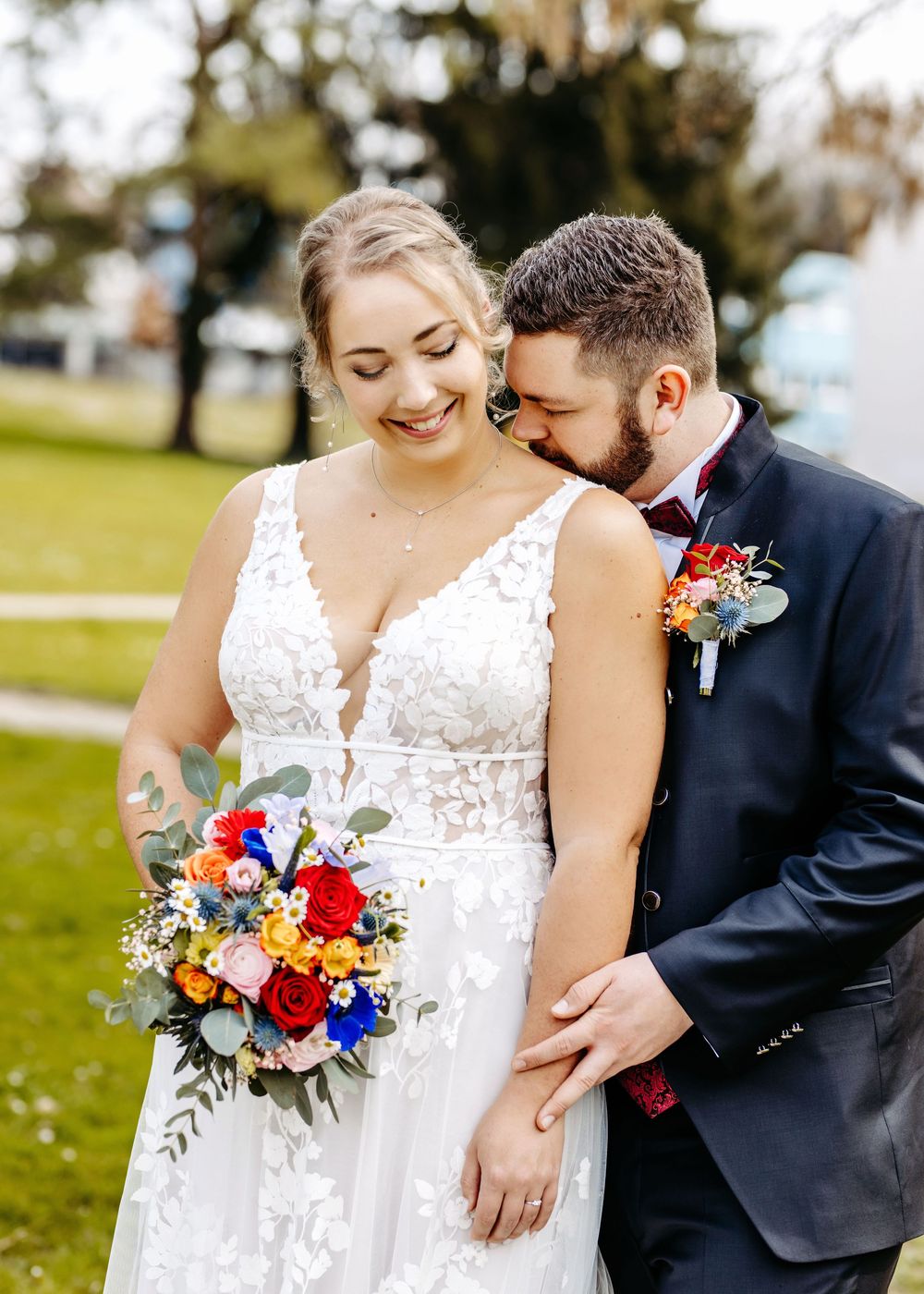 A couple in wedding attire share an intimate moment outdoors with colorful red and blue floral bouquets.