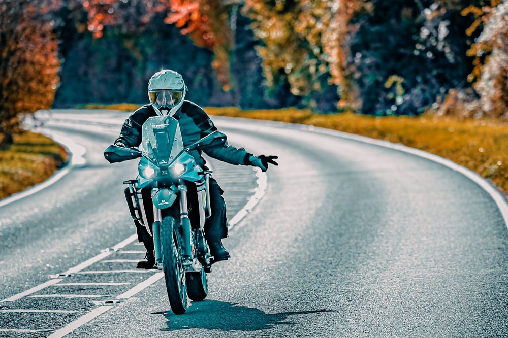 A motorcyclist leans into a curve on a winding mountain road during autumn with colorful foliage in the background.