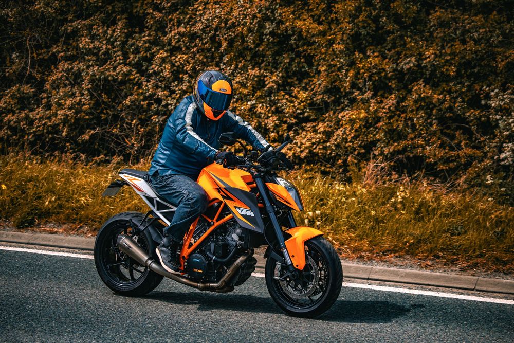 Motorcyclist in blue jacket riding an orange sport bike on a road with autumn foliage in the background.