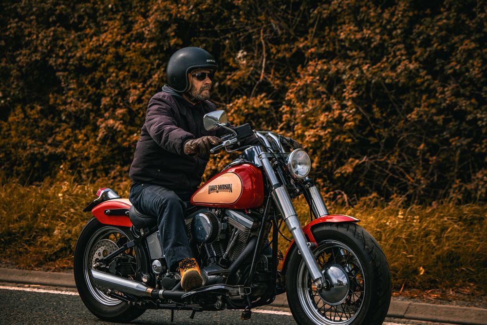 Motorcyclist in dark jacket riding a red cruiser motorcycle on autumn road with orange foliage in background.