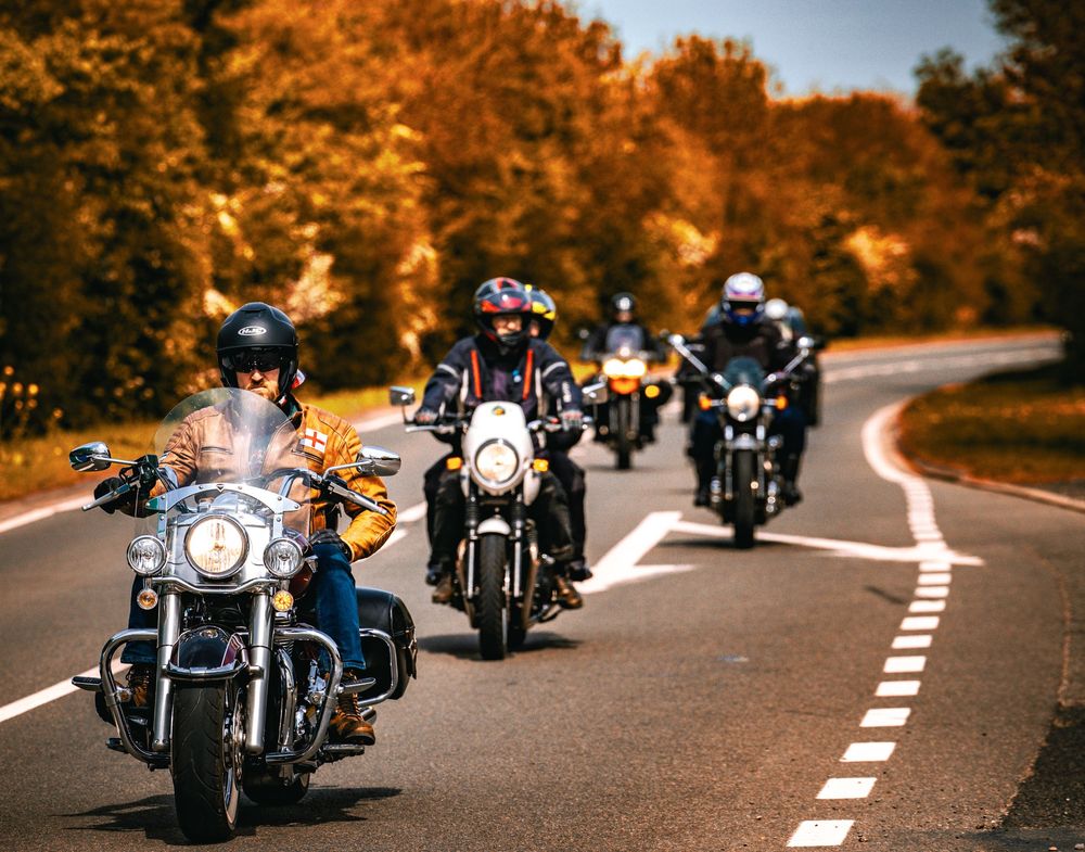 Group of motorcyclists riding cruiser bikes on a curved autumn road lined with orange fall foliage.