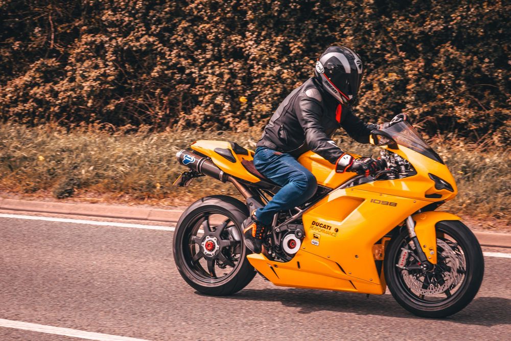 Motorcyclist in black helmet riding yellow sport bike on paved road with autumn foliage in background.