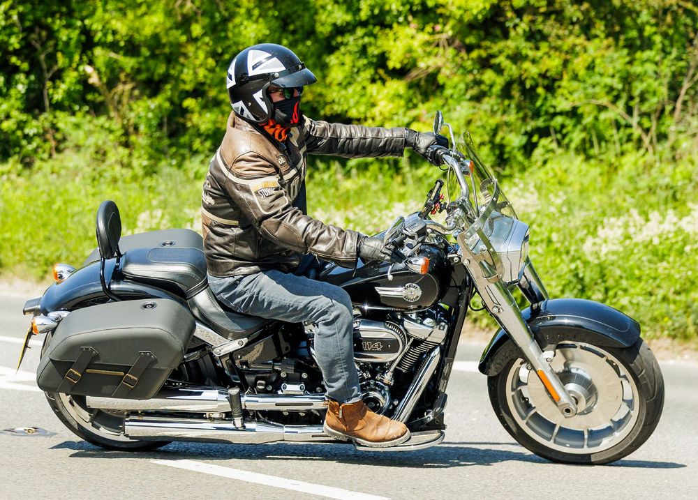 A motorcyclist in protective gear rides a cruiser-style motorcycle on a road with green foliage in the background.