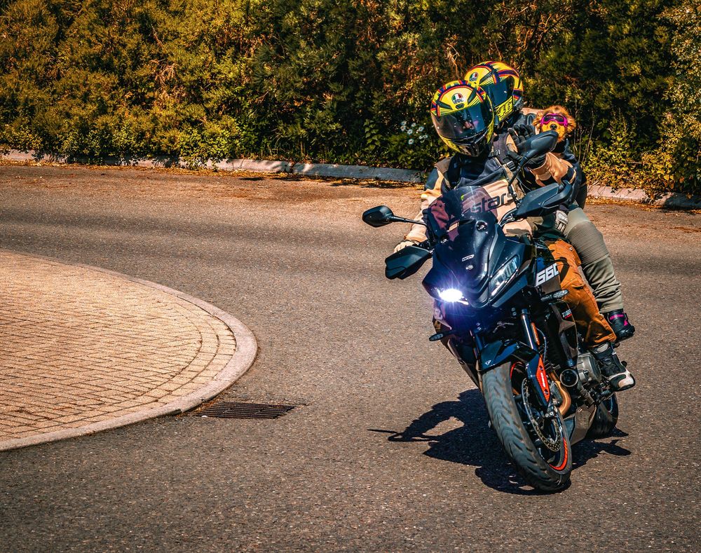 Two motorcyclists riding a blue sport bike on a curved road surrounded by autumn trees.