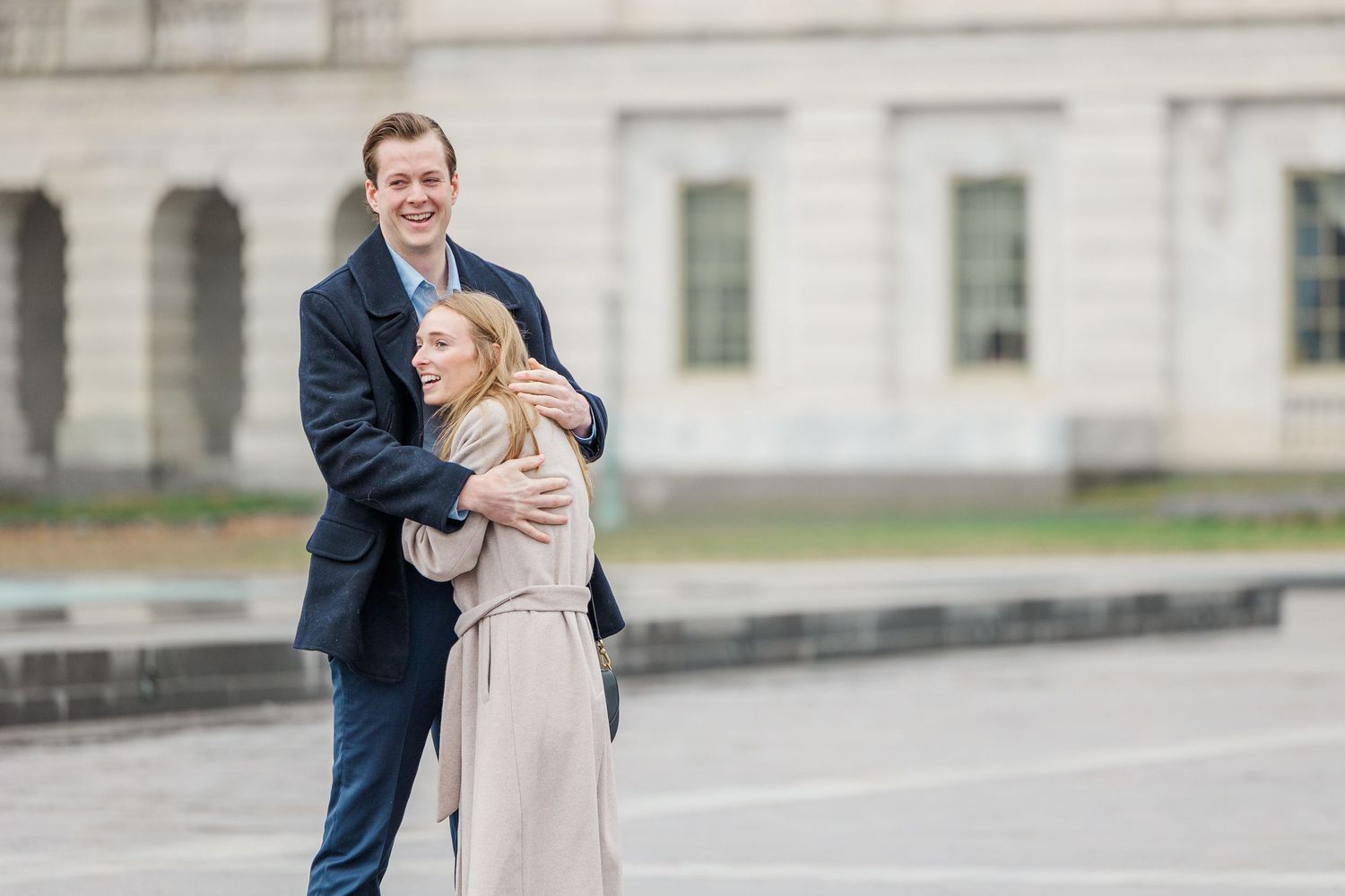 A couple in wedding attire shares a romantic moment on a stone walkway outside a grand building.