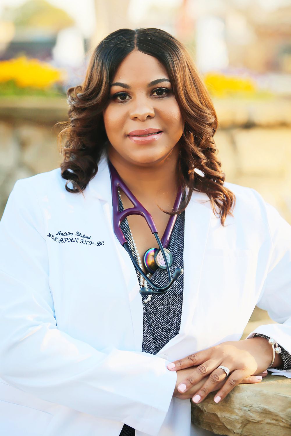 A medical professional in white coat smiles warmly in an outdoor portrait.