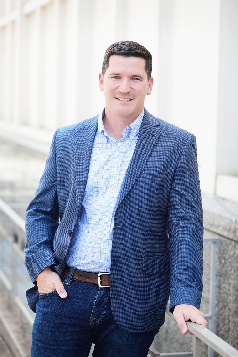 Professional outdoor portrait of a person in a blue blazer leaning on a railing.