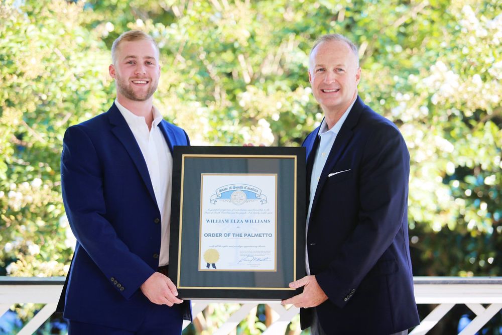 Two men in business attire hold a framed certificate while standing on an outdoor porch.