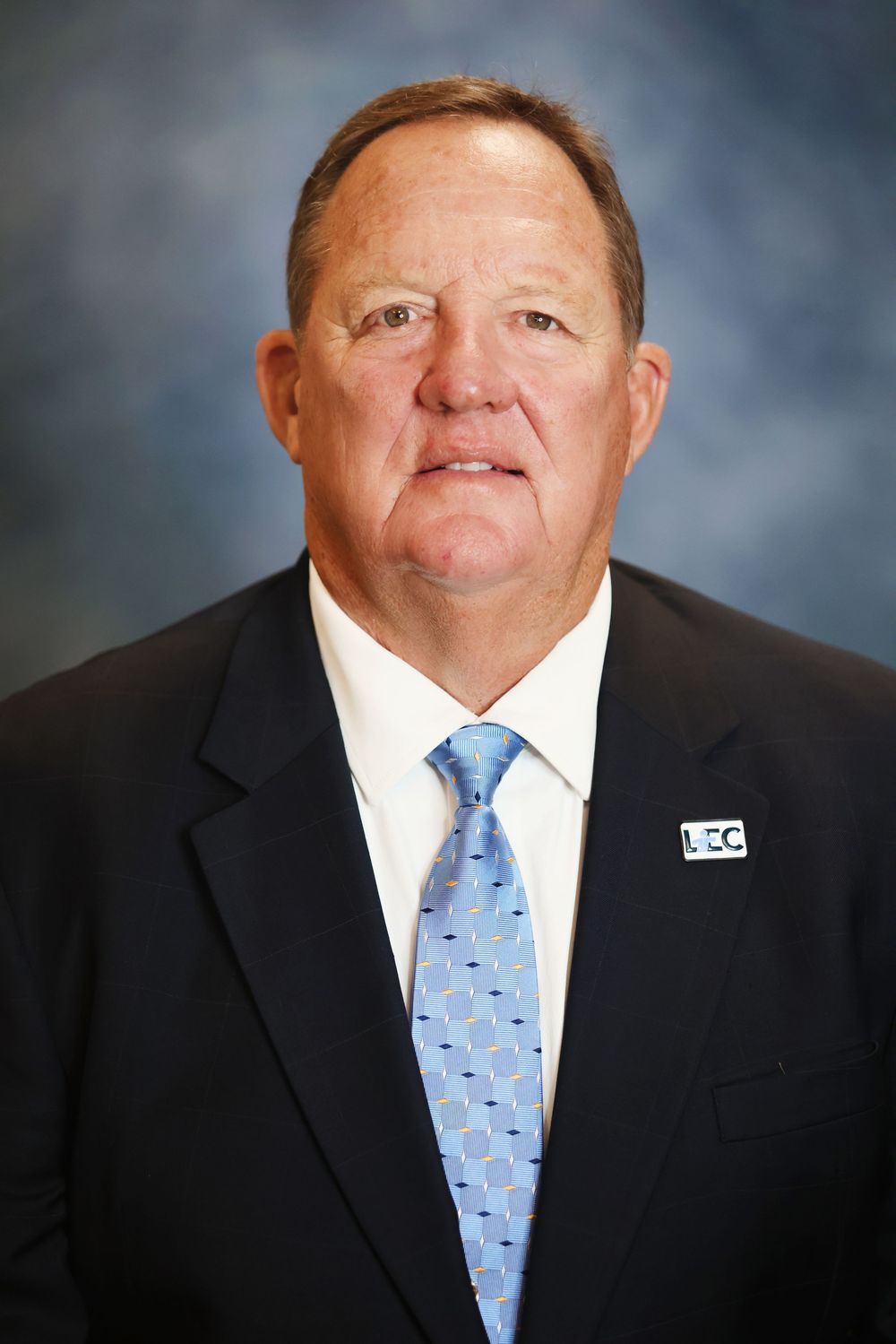 Professional headshot portrait of someone in a dark suit and light blue patterned tie against a mottled gray background.