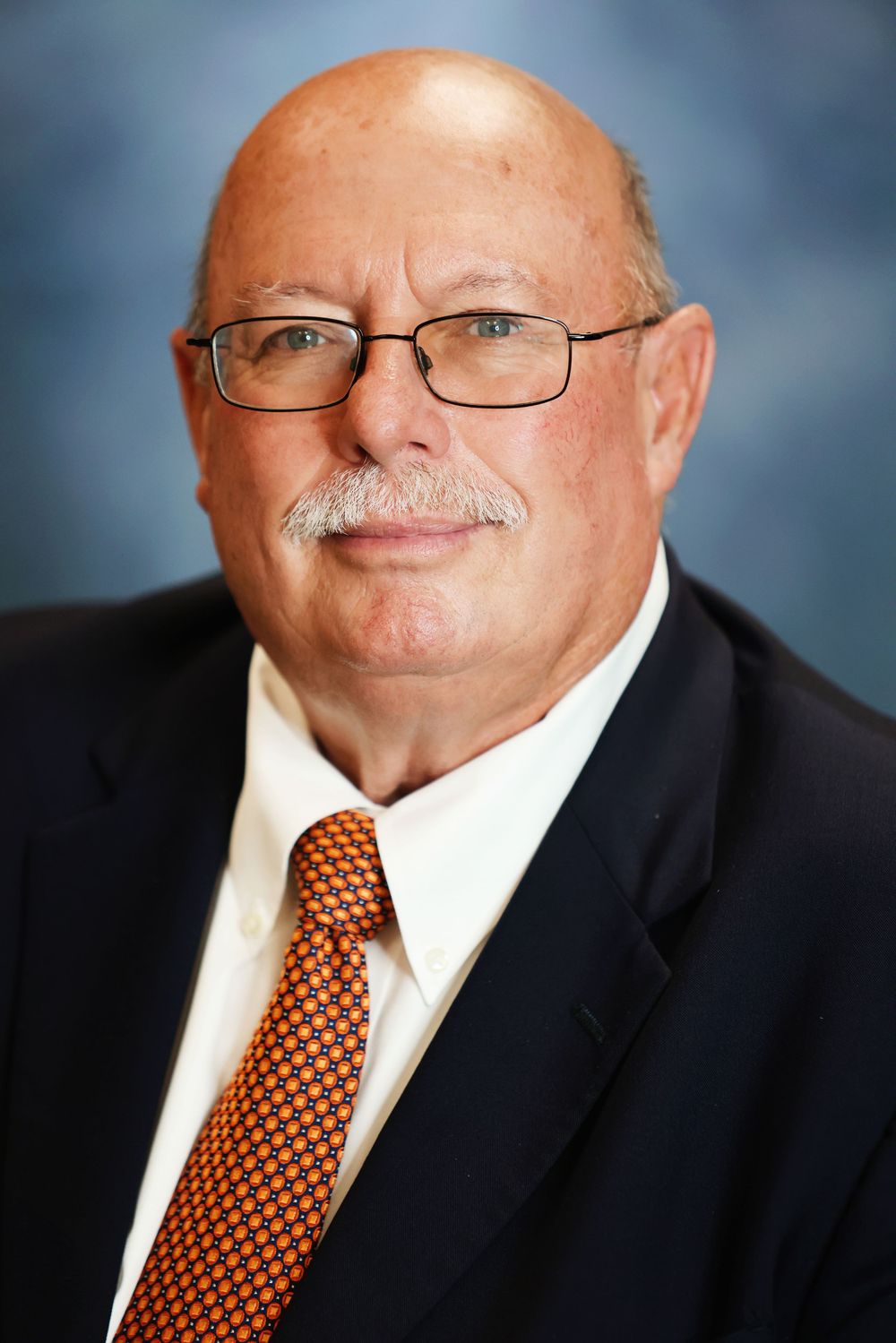 Professional headshot portrait of someone in a black suit and orange polka dot tie against a blue backdrop.