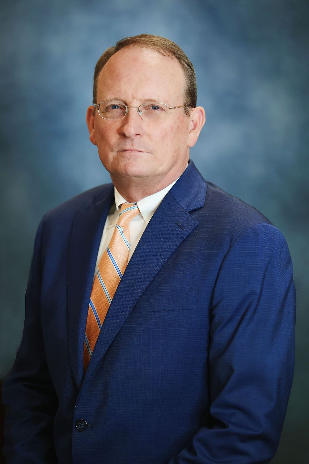 Professional headshot portrait of person in navy suit with orange striped tie against blue studio backdrop.