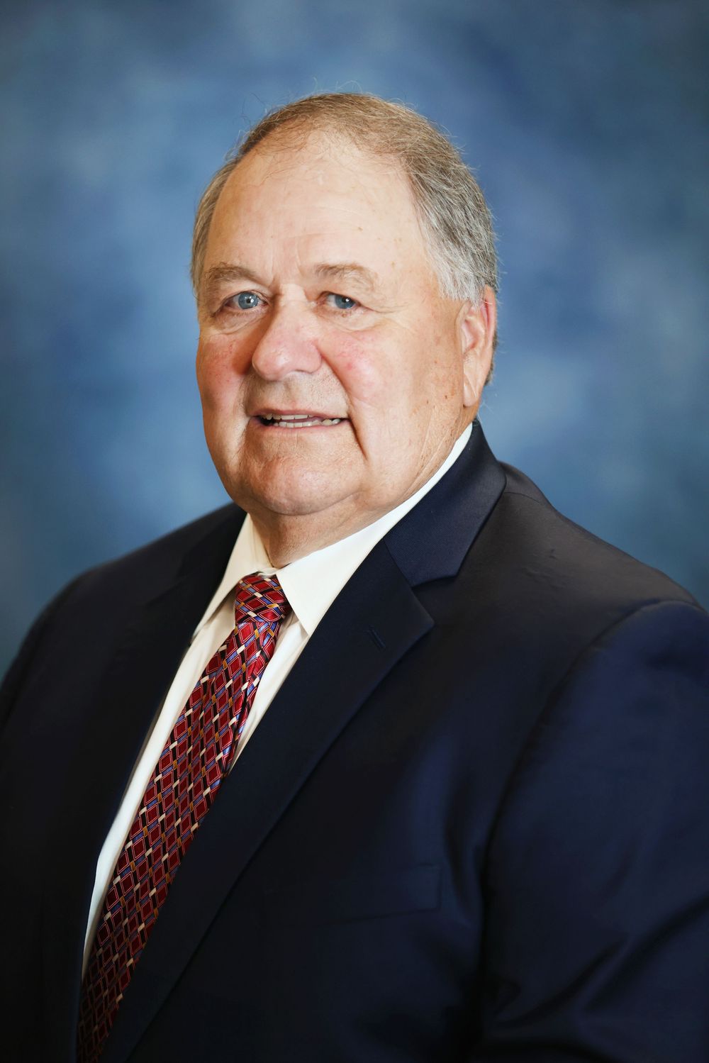 Professional headshot portrait of someone in a navy suit and red patterned tie against a blue mottled background.