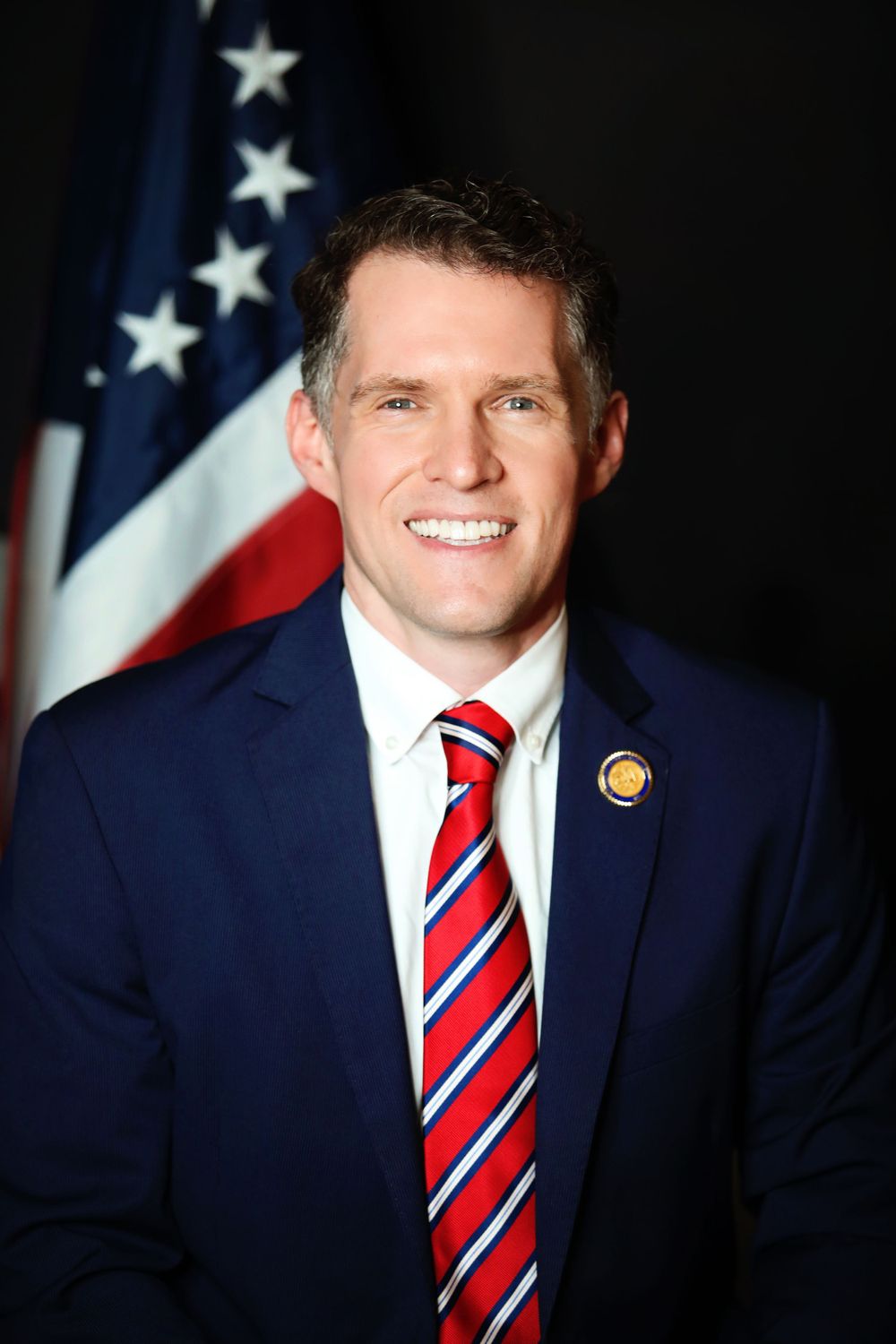 Official portrait of a government representative in navy suit with American flag background.