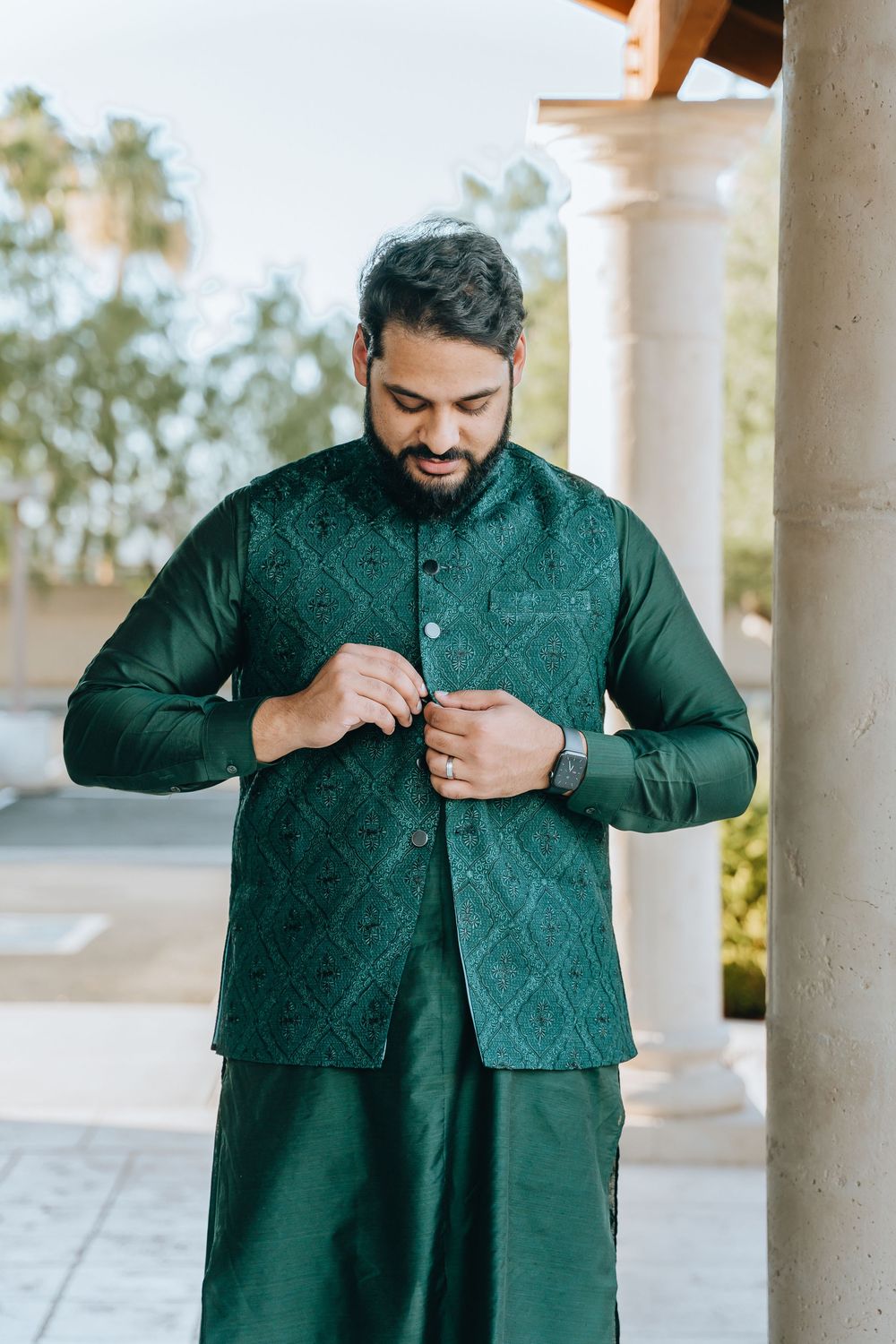 Person in emerald green traditional wear adjusts their outfit in an outdoor colonnade.