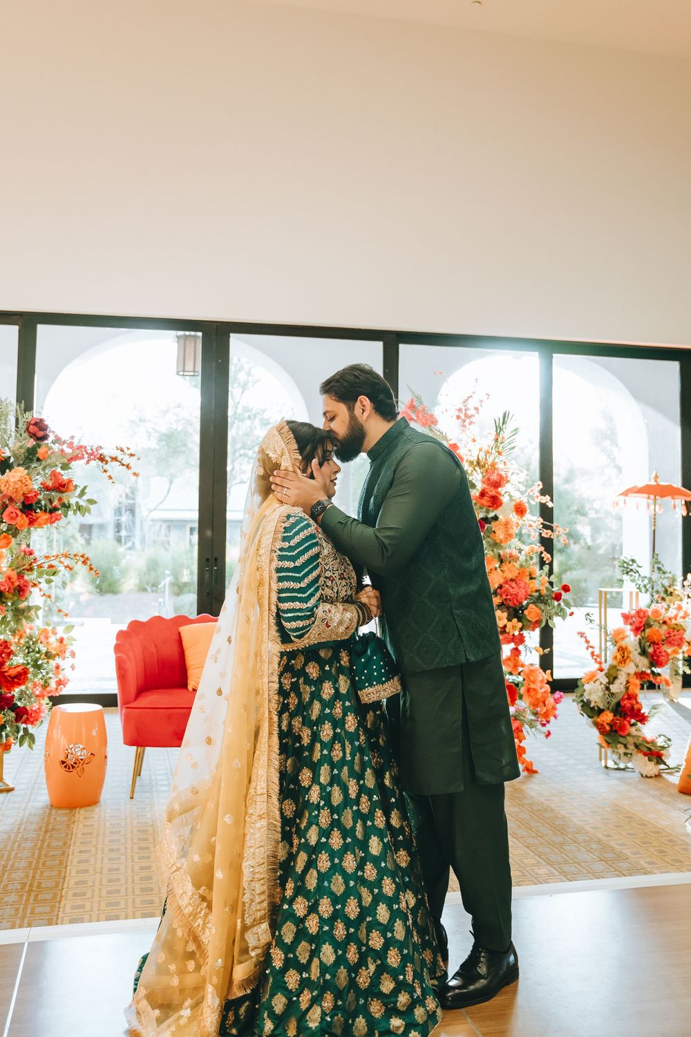Couple in green and gold outfits share an intimate moment in a room with floral decorations.
