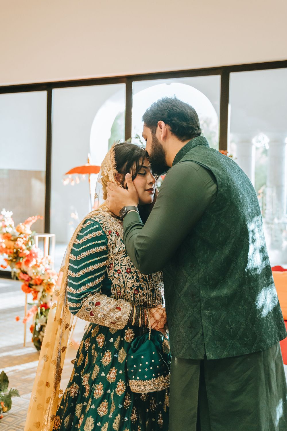 Couple in traditional South Asian wedding attire embrace in an elegantly decorated indoor venue with orange floral accents.