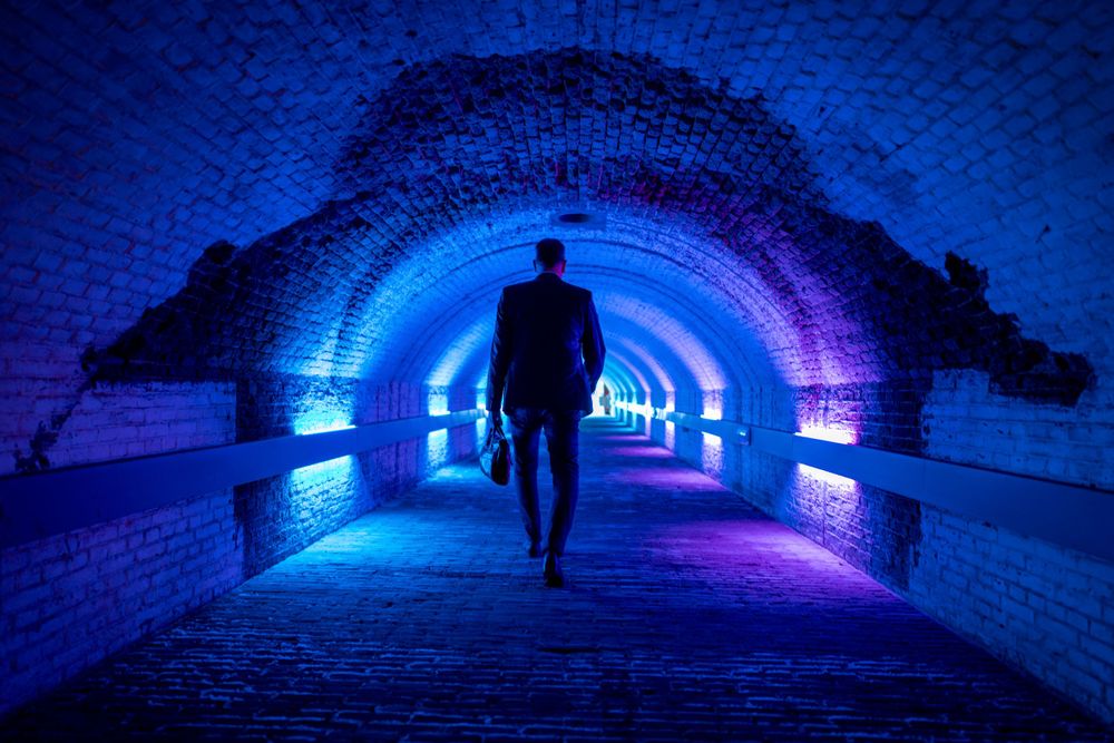 Silhouetted person walking through illuminated tunnel with blue and purple neon lights.