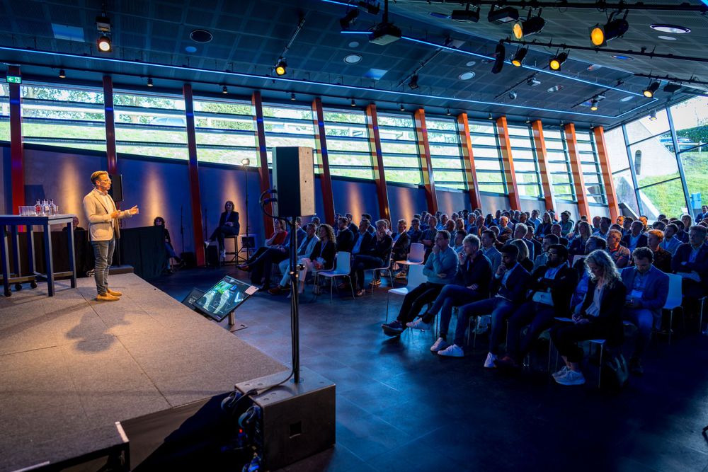 Conference audience seated in dark auditorium with blue lighting and stage.