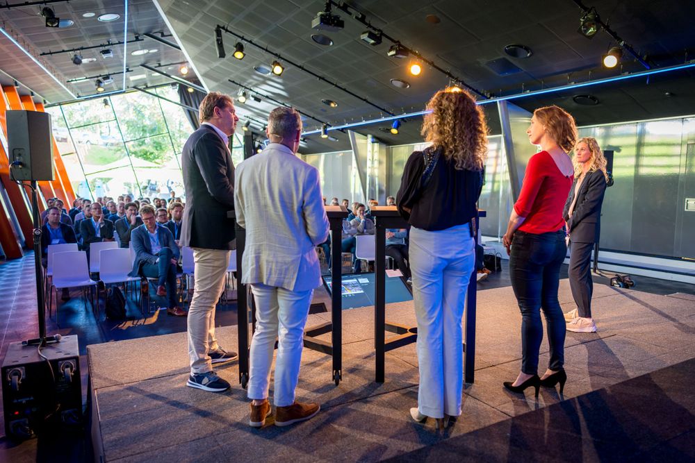 Presenters stand on stage facing an audience during a professional conference or event.
