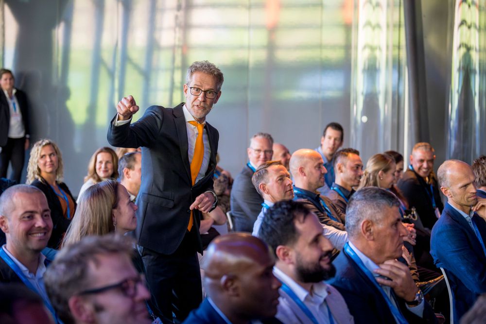 Person in orange tie stands to speak during business conference with audience in foreground.