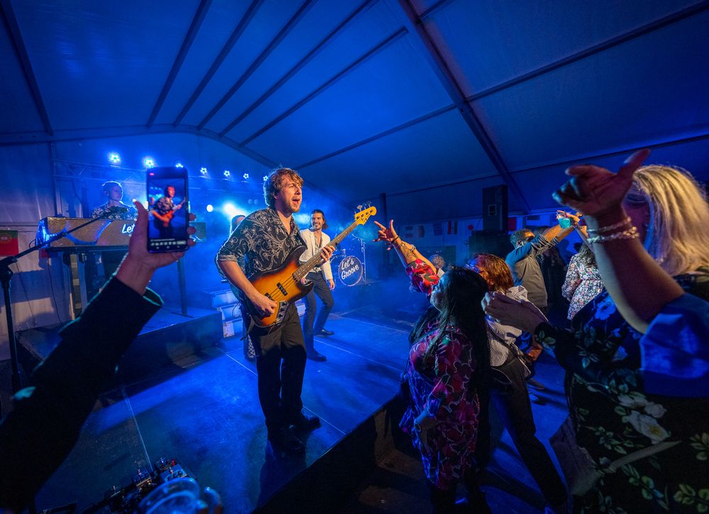 Musicians performing on stage under blue lighting while audience records with phones.