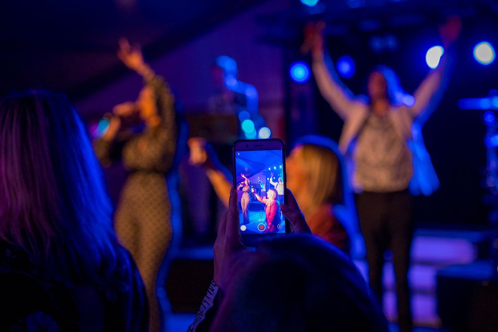 Concert crowd silhouette with person holding up phone to capture performance in blue lighting.