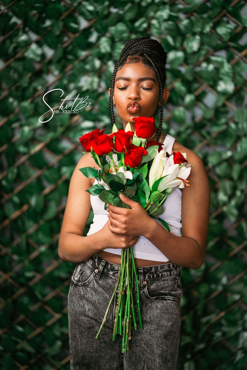 Fashion photo of model in white top and jeans holding red roses against green foliage backdrop.