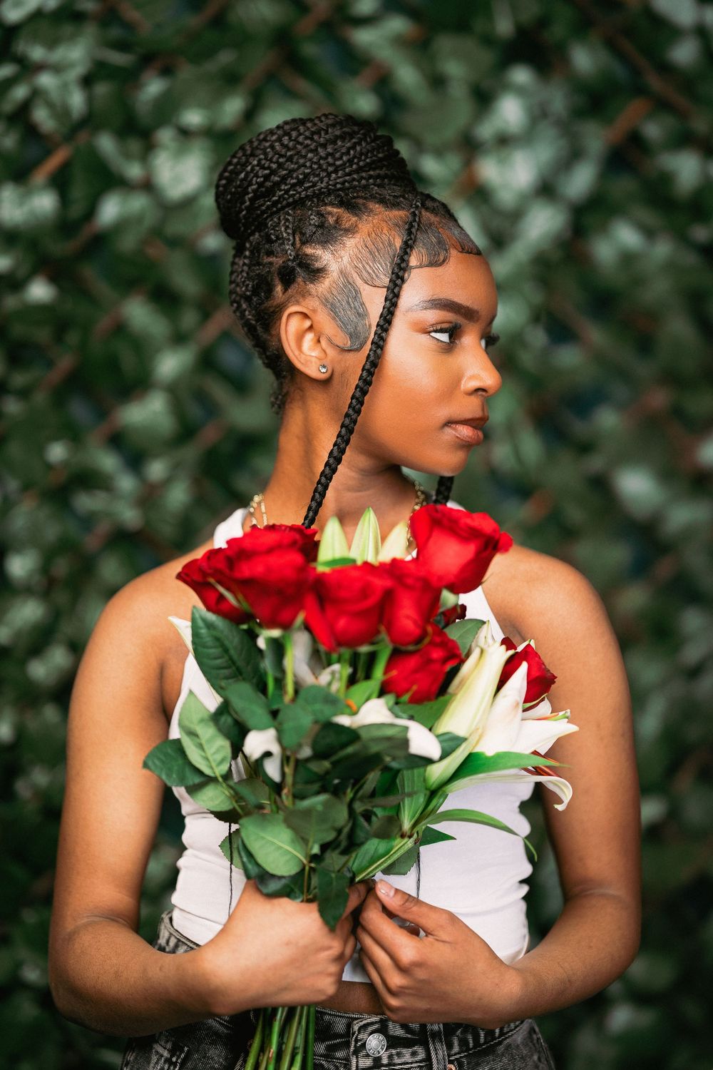 Profile portrait with braided hairstyle holding red roses and white lilies against green foliage.