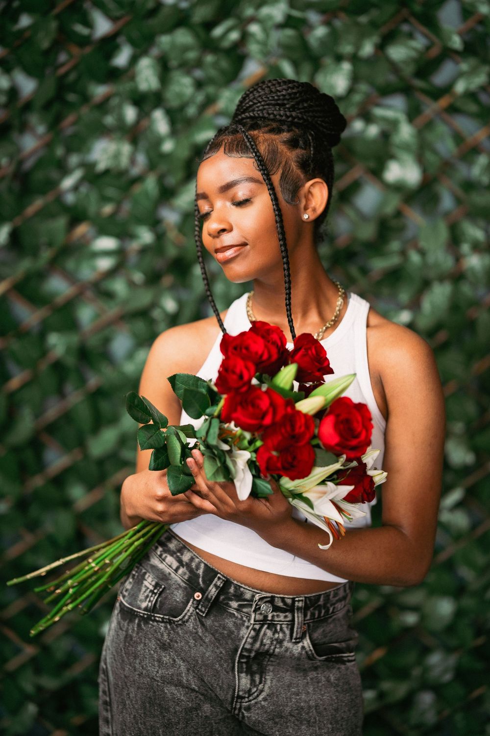 Candid portrait holding red roses with eyes closed against green foliage wearing casual attire.